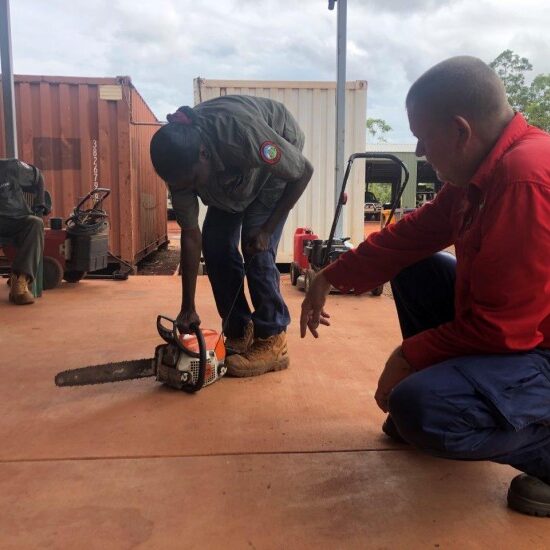 Train Safe NT's Ian van Munster showing Florence Biridjala how to use a chainsaw, with Peter Djigirr looking on